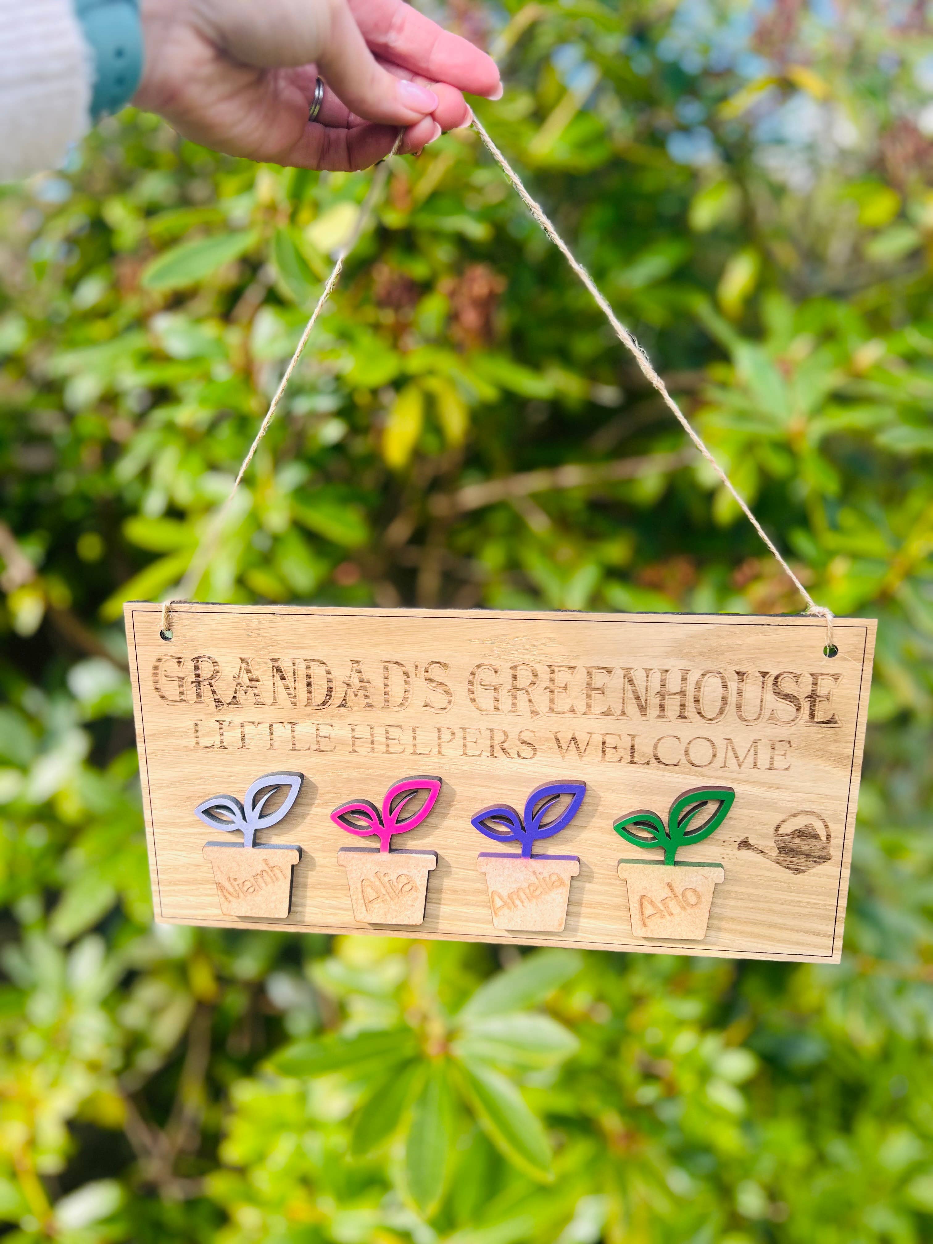 Personalised Grandadโs Greenhouse wooden sign with little helpers welcome text and plant pot design hanging in a GREEN GARDEN.